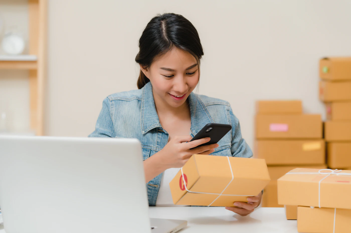 A small business owner checks an order on a phone, beside a laptop and a stack of packed parcels